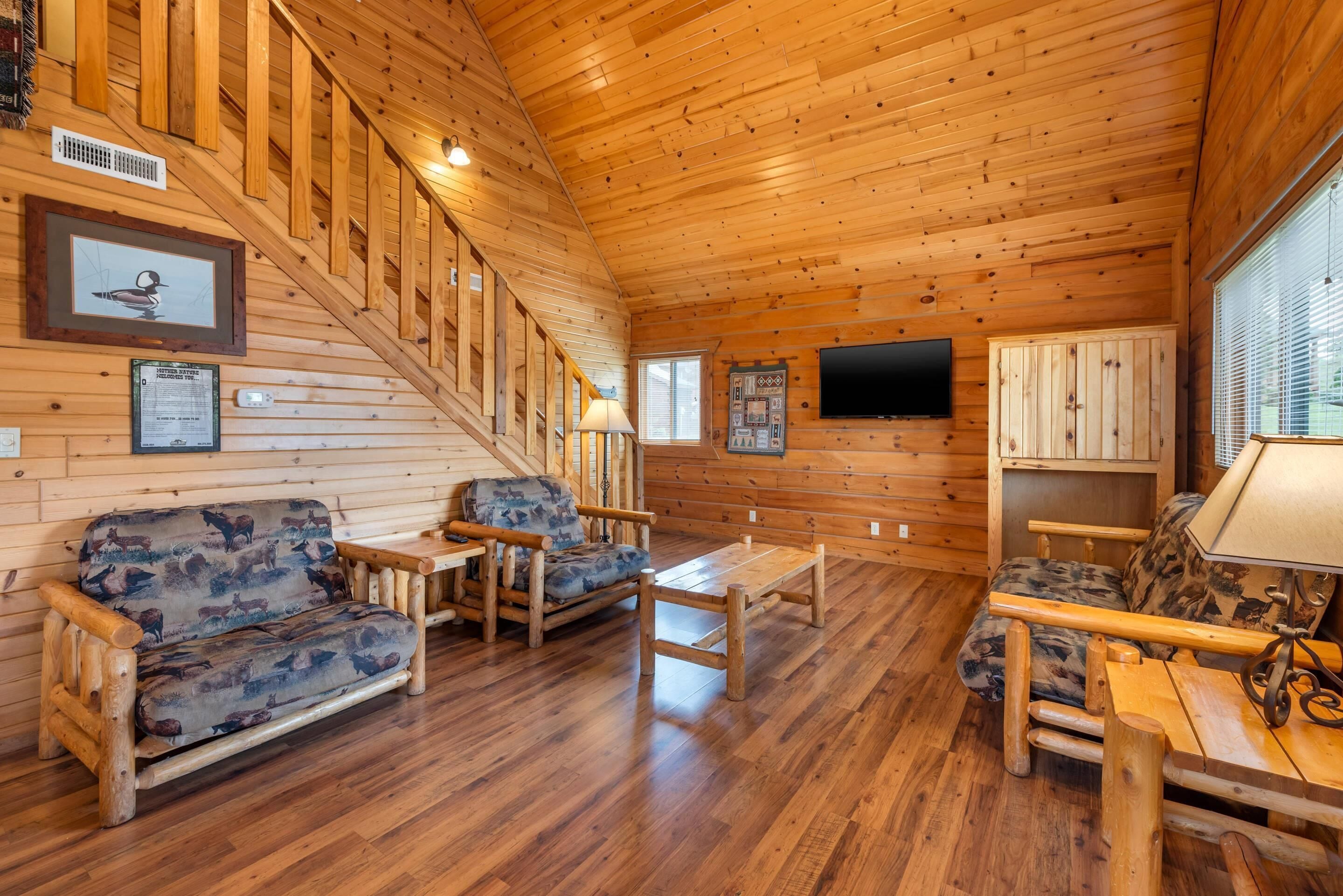 A wooden cabin interior with a sofa, two armchairs, a table, a TV, and a staircase.