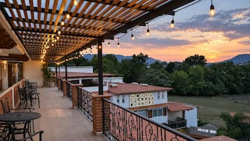 Un hotel con un espacio exterior cubierto que cuenta con mesas y sillas, una pérgola con luces colgantes y vistas del paisaje circundante.
