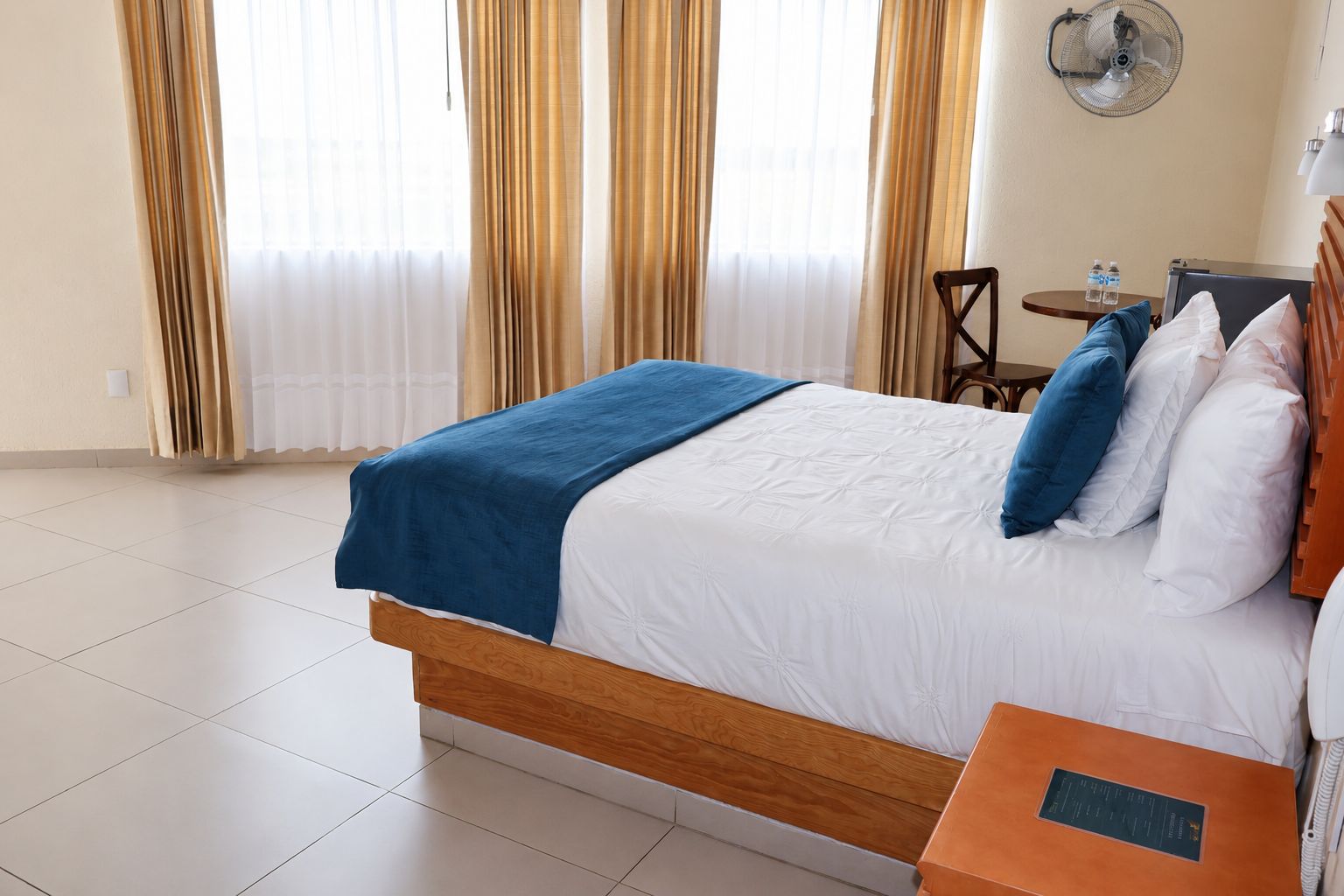 Una habitación de hotel con cama, mesita de noche, silla, ventilador y cortinas.