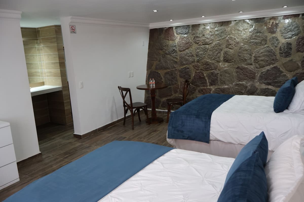 Habitación de hotel con dos camas, una mesita pequeña, una silla, un muro decorativo de piedra y zona de ducha.