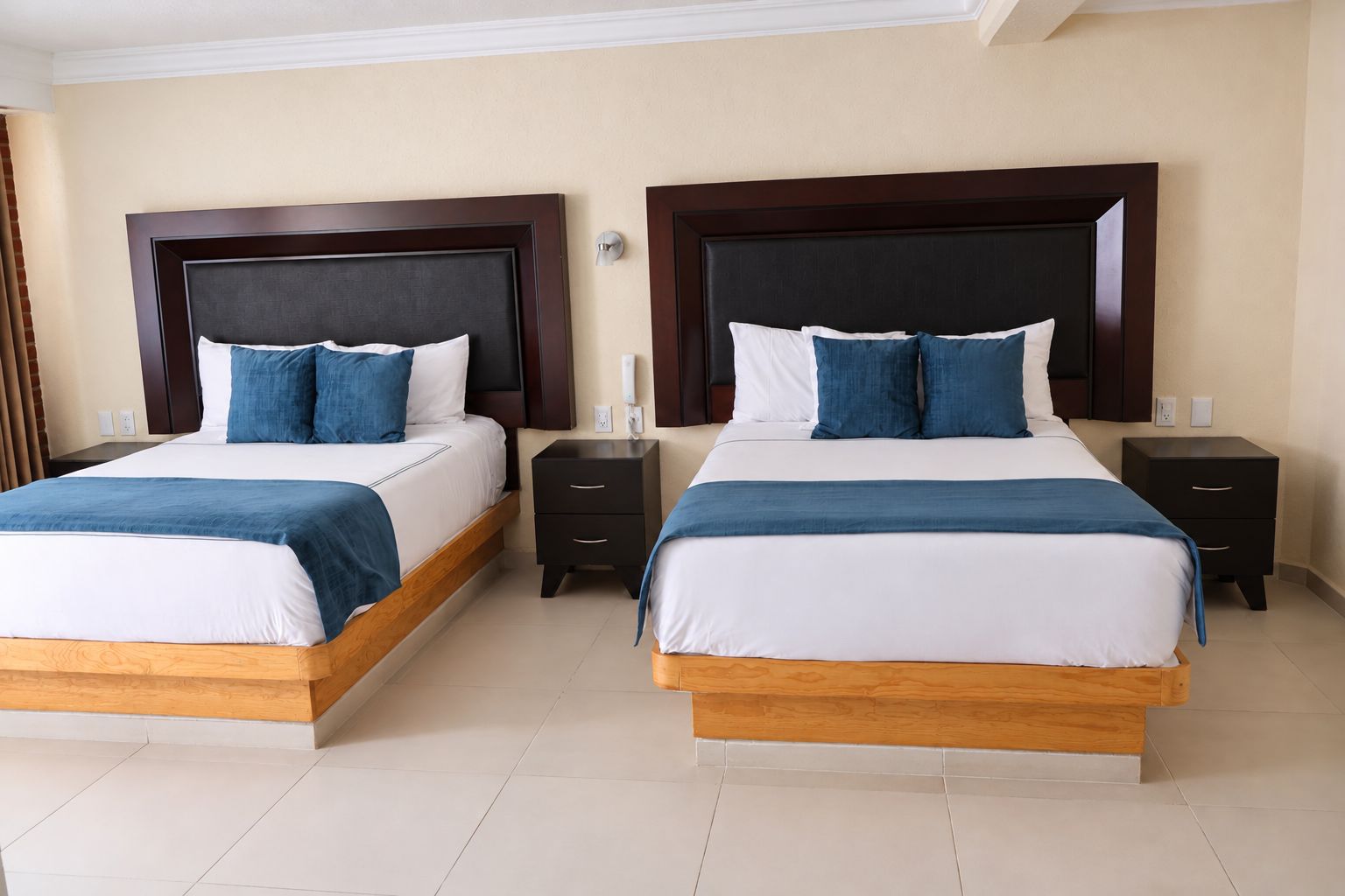 A hotel room with two beds, each with a wooden headboard, white bedding, and blue throw pillows. There are bedside tables with drawers and a wall-mounted light fixture.