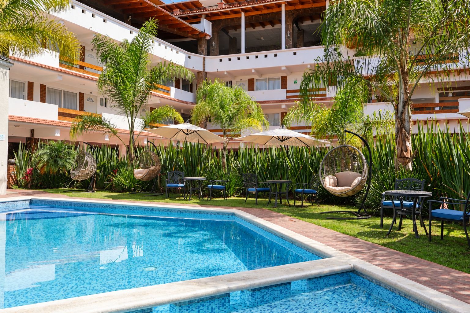 A hotel pool area with a clear blue pool, surrounded by lush greenery and outdoor seating.