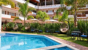 A hotel pool area with a clear blue pool, surrounded by lush greenery and outdoor seating.