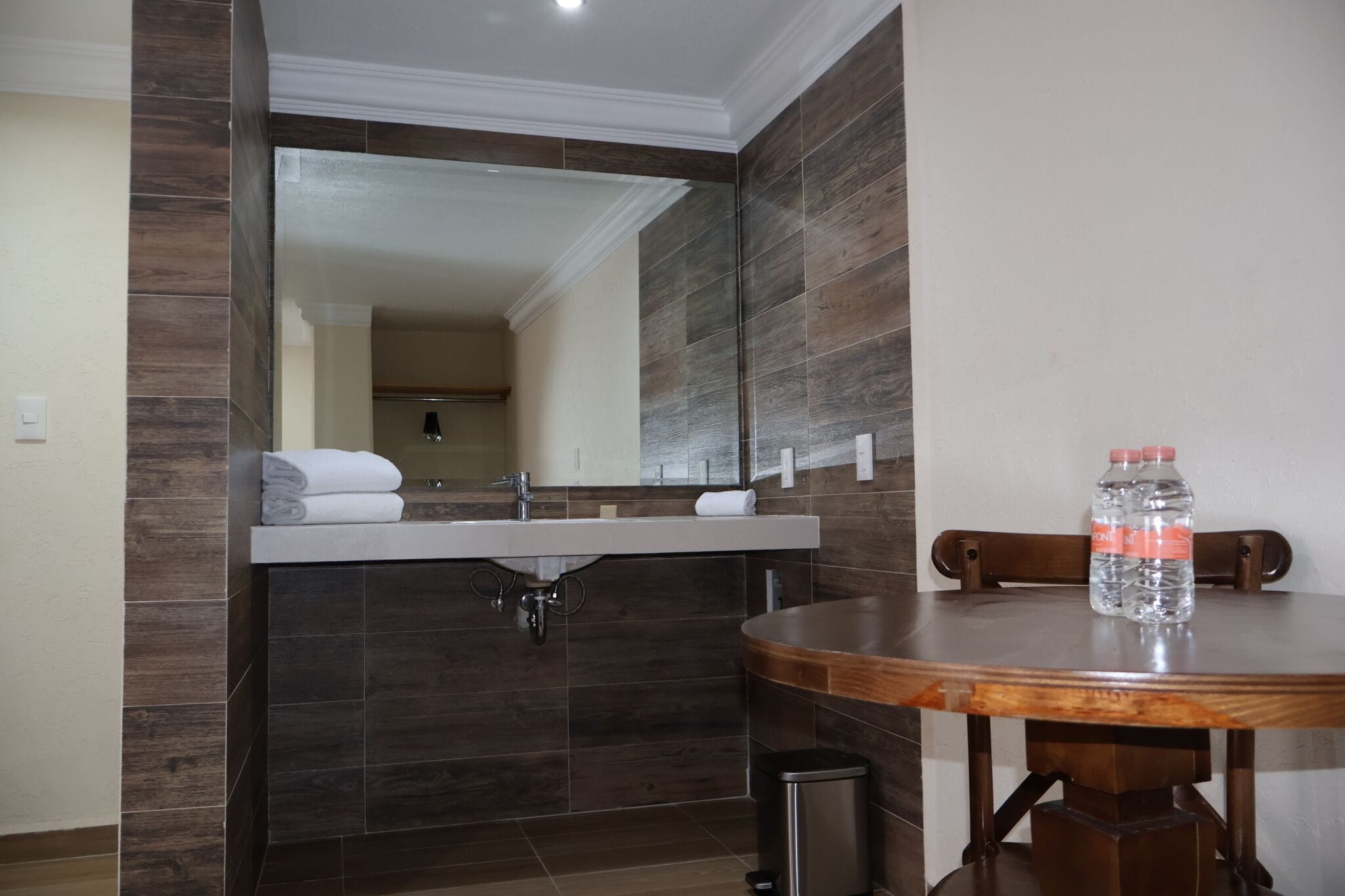 Une salle de bain équipée d’un lavabo, d’un miroir et d’une table ronde sur laquelle sont posées deux bouteilles d’eau.