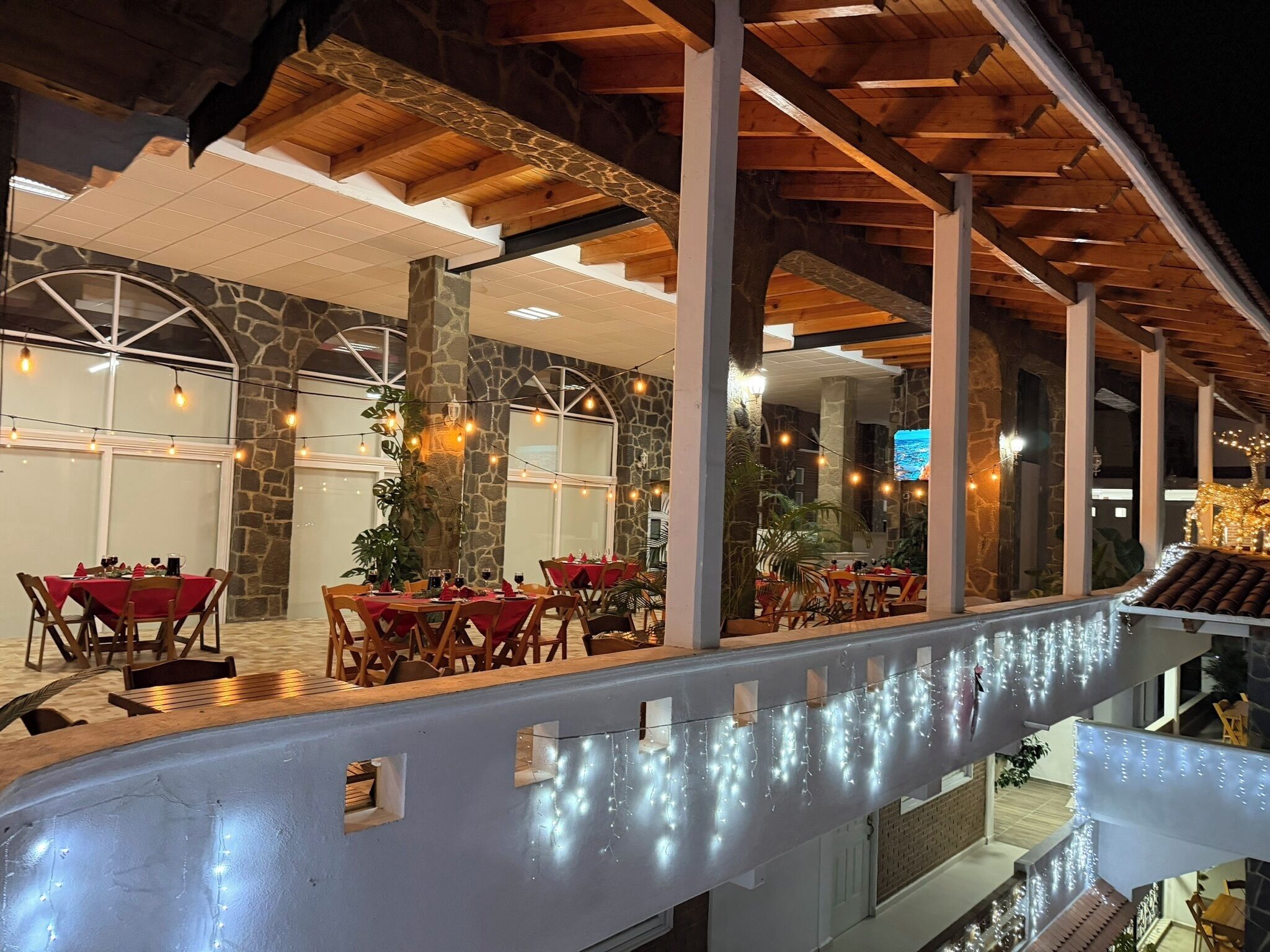 Un espace de restauration intérieur aux murs en pierre, aux poutres en bois et aux guirlandes lumineuses. Les tables sont dressées avec des nappes rouges et des chaises.