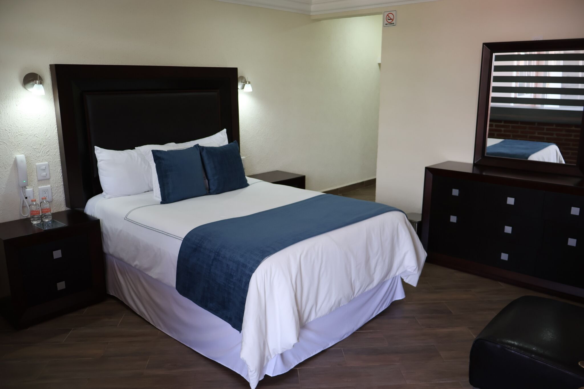 A neatly made bed with a dark headboard, white and blue bedding, a small bedside table with a bottle and glass, and a dresser with a mirror.