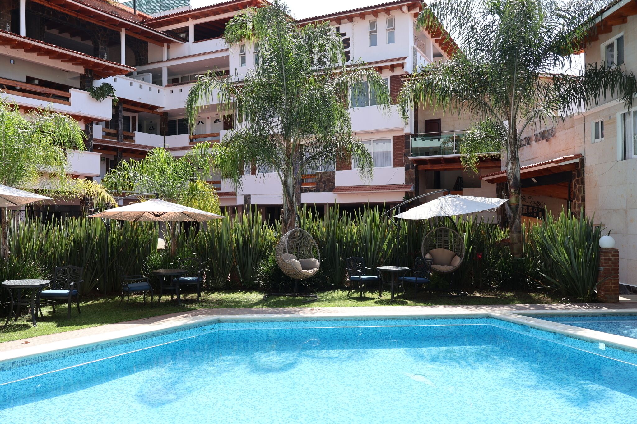 A pool area with a clear blue pool, surrounded by lounge chairs and umbrellas, and a multi-story building with balconies in the background.