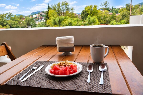 Mesa de madeira com prato de comida, xícara de café e copo d'água em varanda com vista para área verde e edifícios.