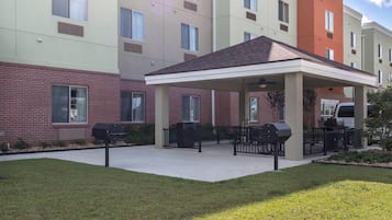A covered outdoor patio with barbecue grills and seating, adjacent to a multi-story building with a brick facade and multiple windows.
