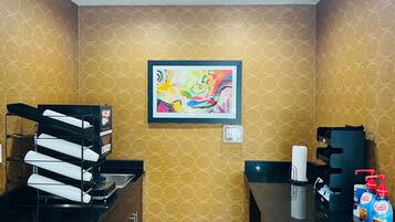 Una habitación de hotel con papel tapiz estampado, un cuadro abstracto enmarcado, un escritorio negro con cafetera y una mesa con dispensadores de café.