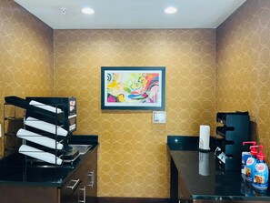 Una habitación de hotel con papel tapiz estampado, un cuadro abstracto enmarcado, un escritorio negro con cafetera y una mesa con dispensadores de café.