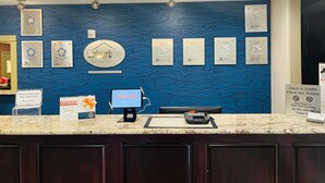 A hotel reception area with a blue wavy wall, framed awards, a digital welcome screen, and a check-in counter. - Suburban Studios Donaldsonville - Gonzales (Donaldsonville)
