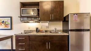 A compact kitchen with dark wooden cabinets, a stainless steel refrigerator, a microwave, and a granite countertop. - Suburban Studios Donaldsonville - Gonzales (Donaldsonville)