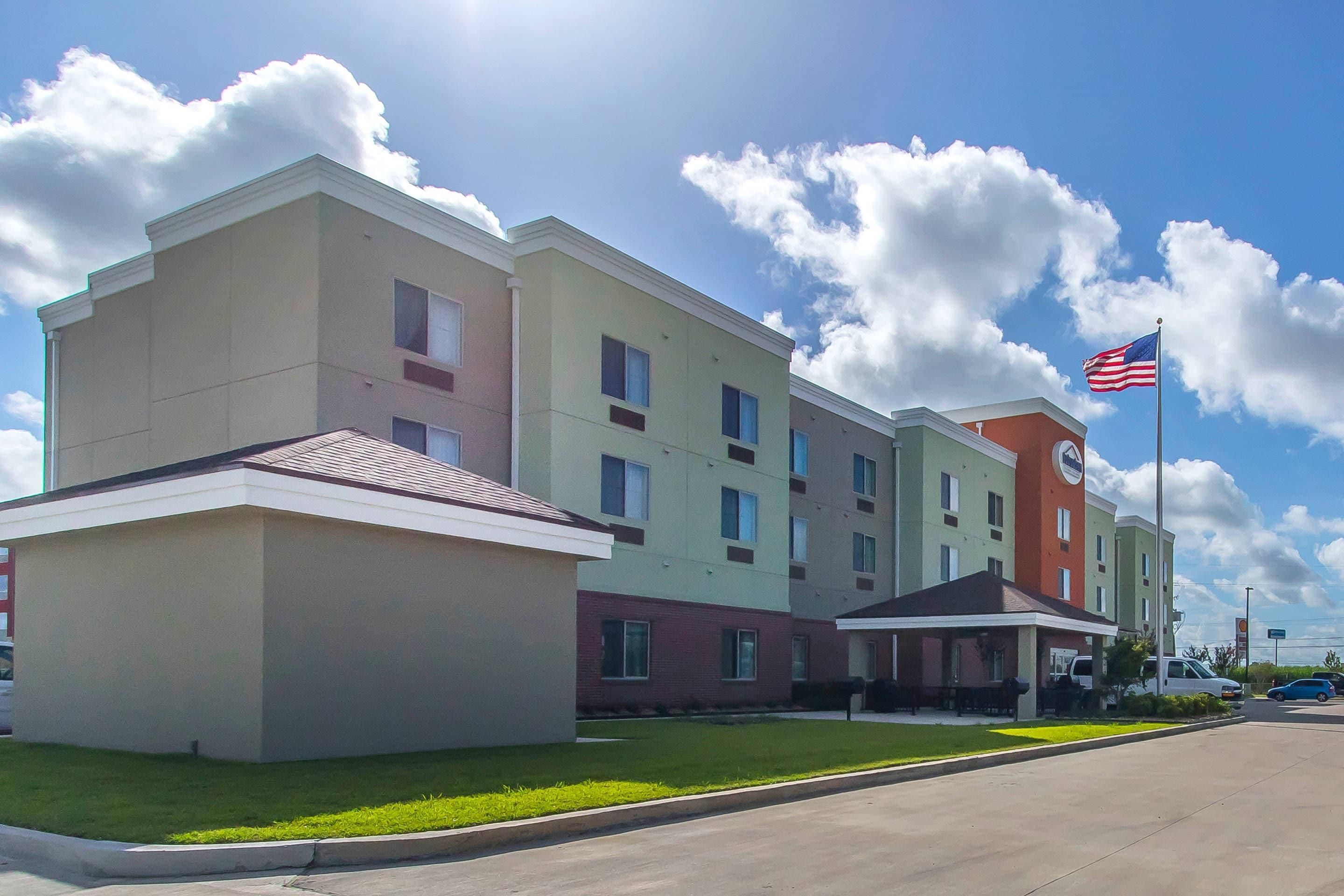 A multi-story hotel with a flat roof, a flagpole with an American flag, and a parking lot with cars.