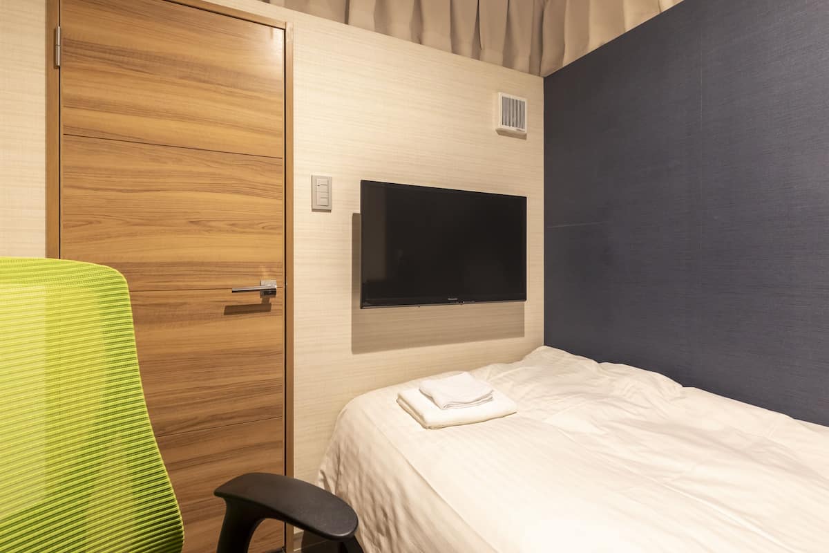 A hotel room with a wooden wardrobe, a green chair, a bed with a white comforter, and a mounted flat-screen TV.