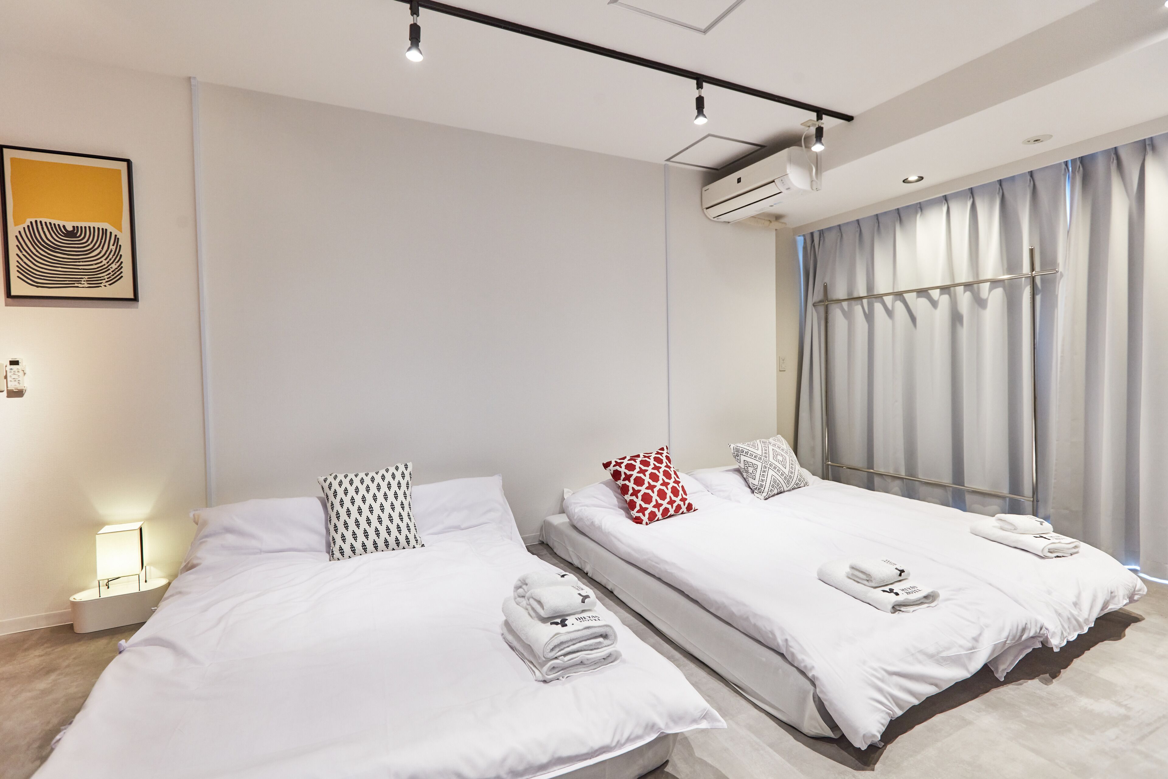A modern hotel room with two beds, a bedside table with a lamp, a large window with curtains, and an air conditioning unit.