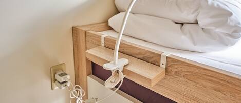 Una cama con manta y almohadas blancas, cabecera de madera y lámpara de noche blanca.