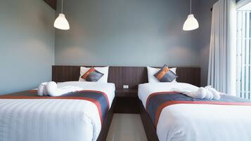 A hotel room with two beds, each with a headboard, white bedding, and orange pillows. There are two pendant lights hanging from the ceiling, and a window with curtains on the right side.
