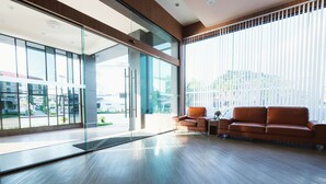 A modern lobby with a wooden floor, a leather sofa, and large windows offering a view of the outside. - Hansanan Hotel (Phitsanulok)