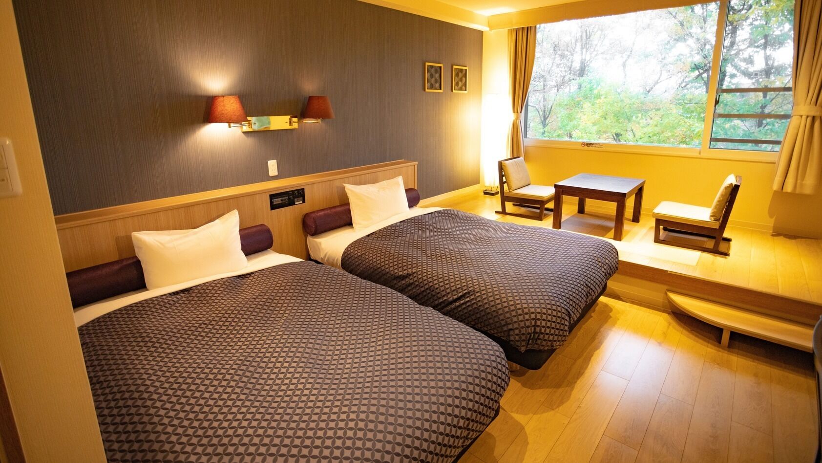 Habitación de hotel con dos camas, una mesita, una silla y un ventanal con vistas a árboles.