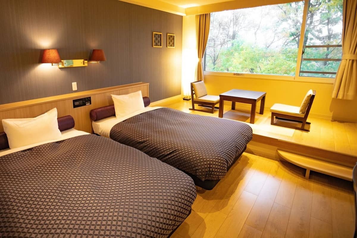 Hotellihuone, jossa on kaksi sänkyä, pieni pöytä, tuoli ja ikkuna, josta on näkymä puita kohti.