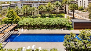Une piscine dotée d’une terrasse en bois et de chaises longues, entourée de palmiers et de verdure, avec des bâtiments modernes en arrière-plan.