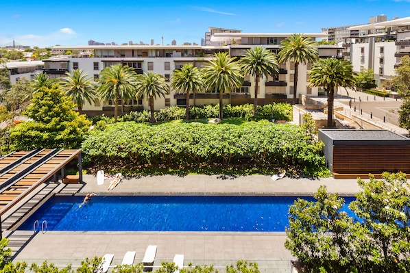 A swimming pool with a wooden deck and lounge chairs, surrounded by palm trees and greenery, with modern buildings in the background. - Stylish 1 Bedroom Apartment Camperdown (Camperdown)