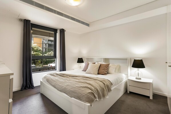 Een moderne slaapkamer met een groot bed, nachtkastjes, een raam met gordijnen en uitzicht op gebouwen.