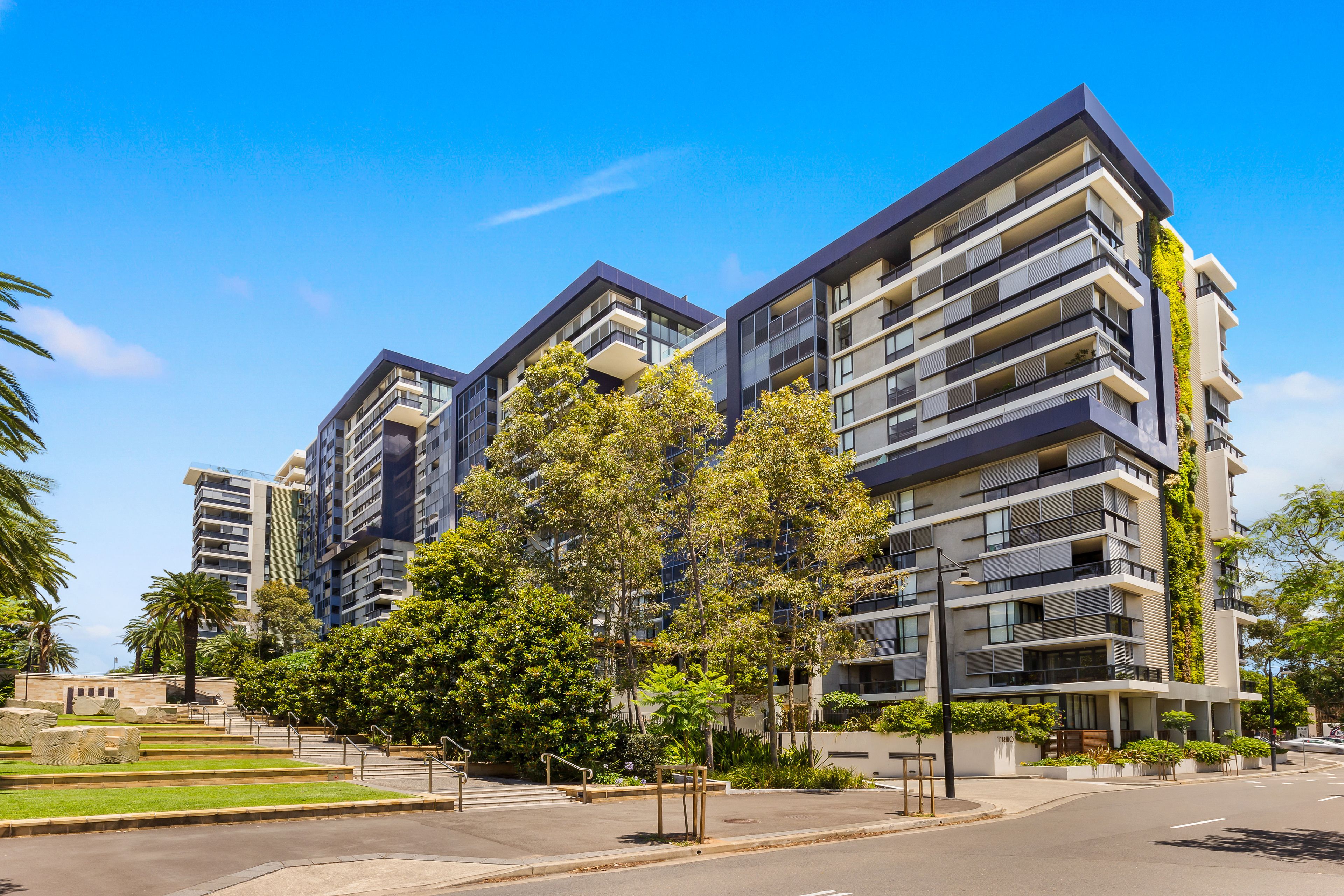 A modern multi-story residential building with balconies, surrounded by greenery and a clear sky.