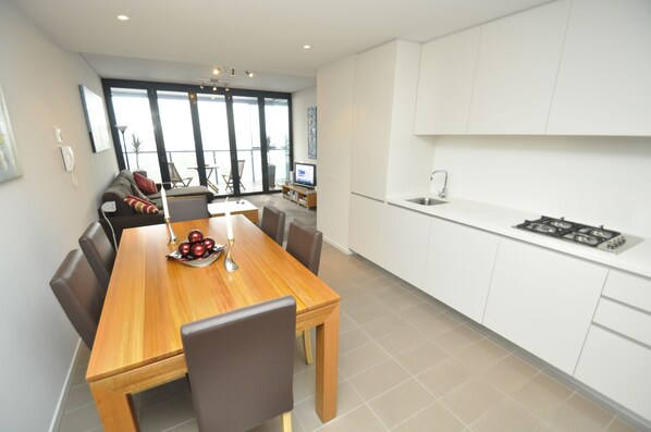 A modern kitchen with white cabinets, a wooden dining table, and a large window overlooking a balcony. - Stylish 1 Bedroom Apartment Camperdown (Camperdown)
