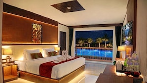 A modern hotel room with a large bed, a view of a swimming pool at night, and a balcony with palm trees.