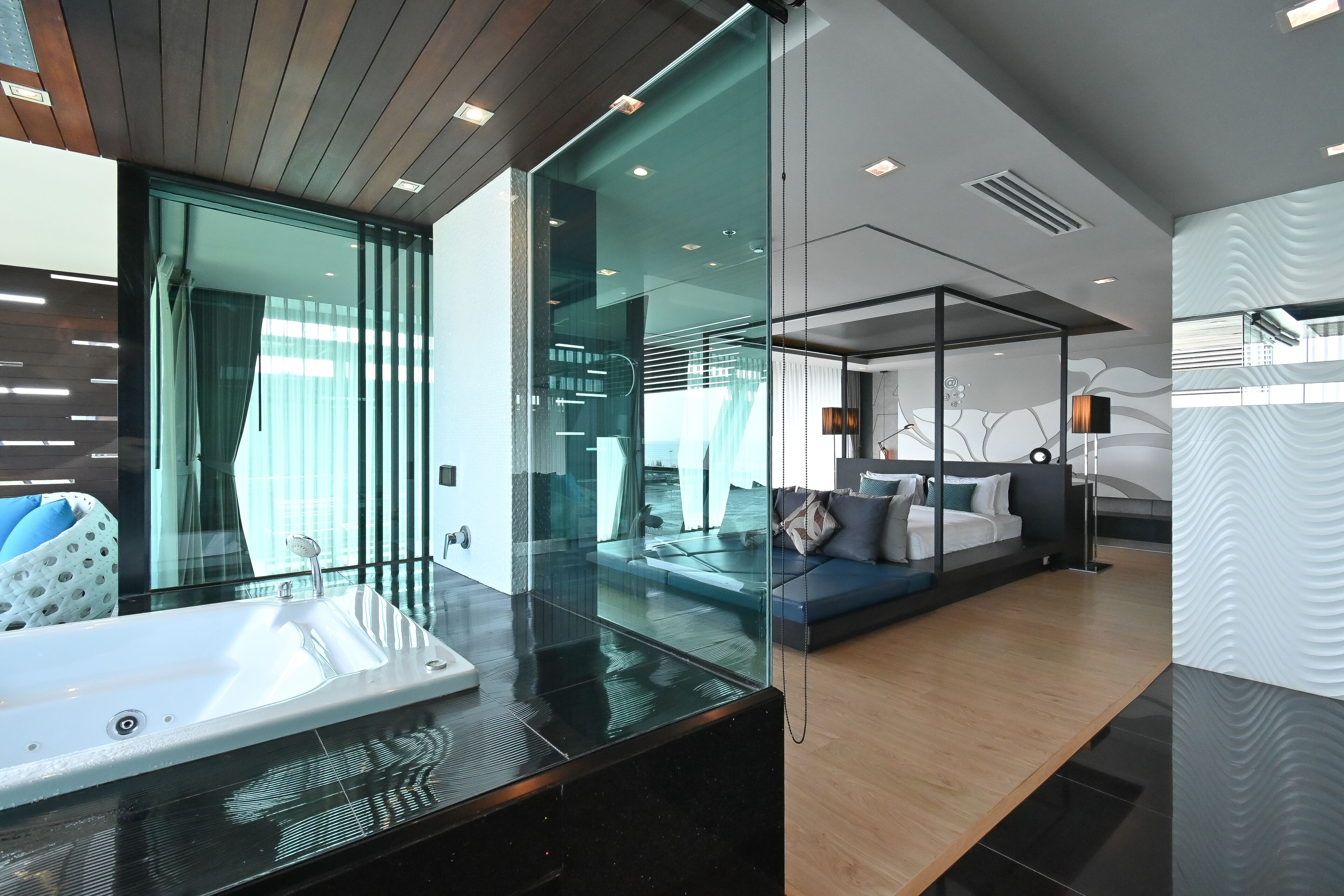 A modern hotel room with a glass partition, a large white bathtub, a sofa with cushions, and a wooden floor.
