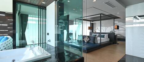 A modern hotel room with a glass partition, a large white bathtub, a sofa with cushions, and a wooden floor.