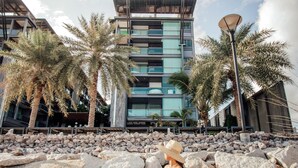 A modern building with a stone facade, palm trees, and a person sitting on rocks.