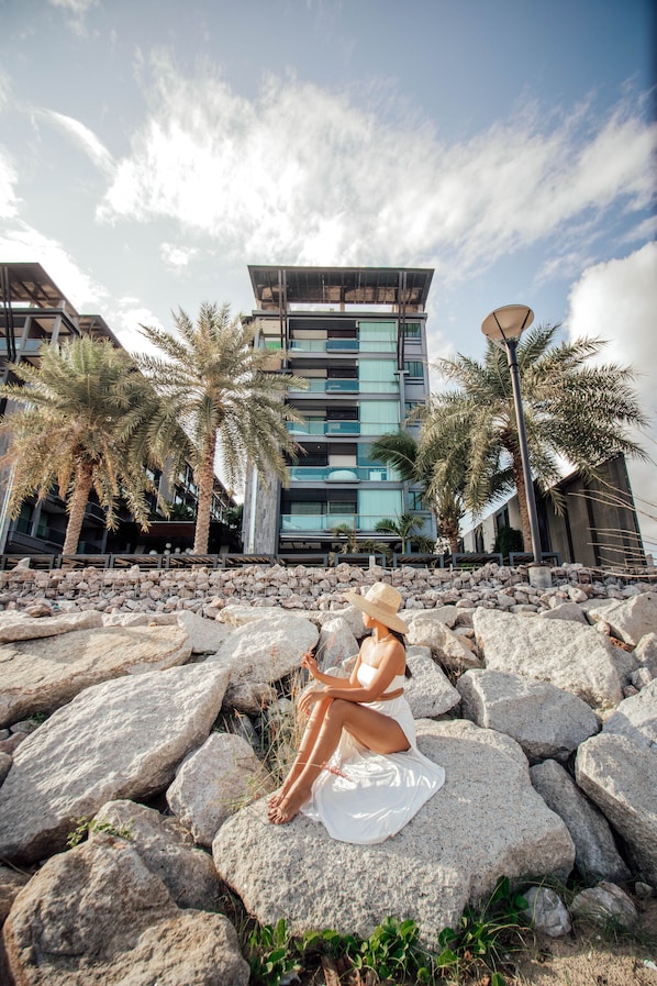 Un edificio moderno con fachada de piedra, palmeras y una persona sentada sobre rocas.