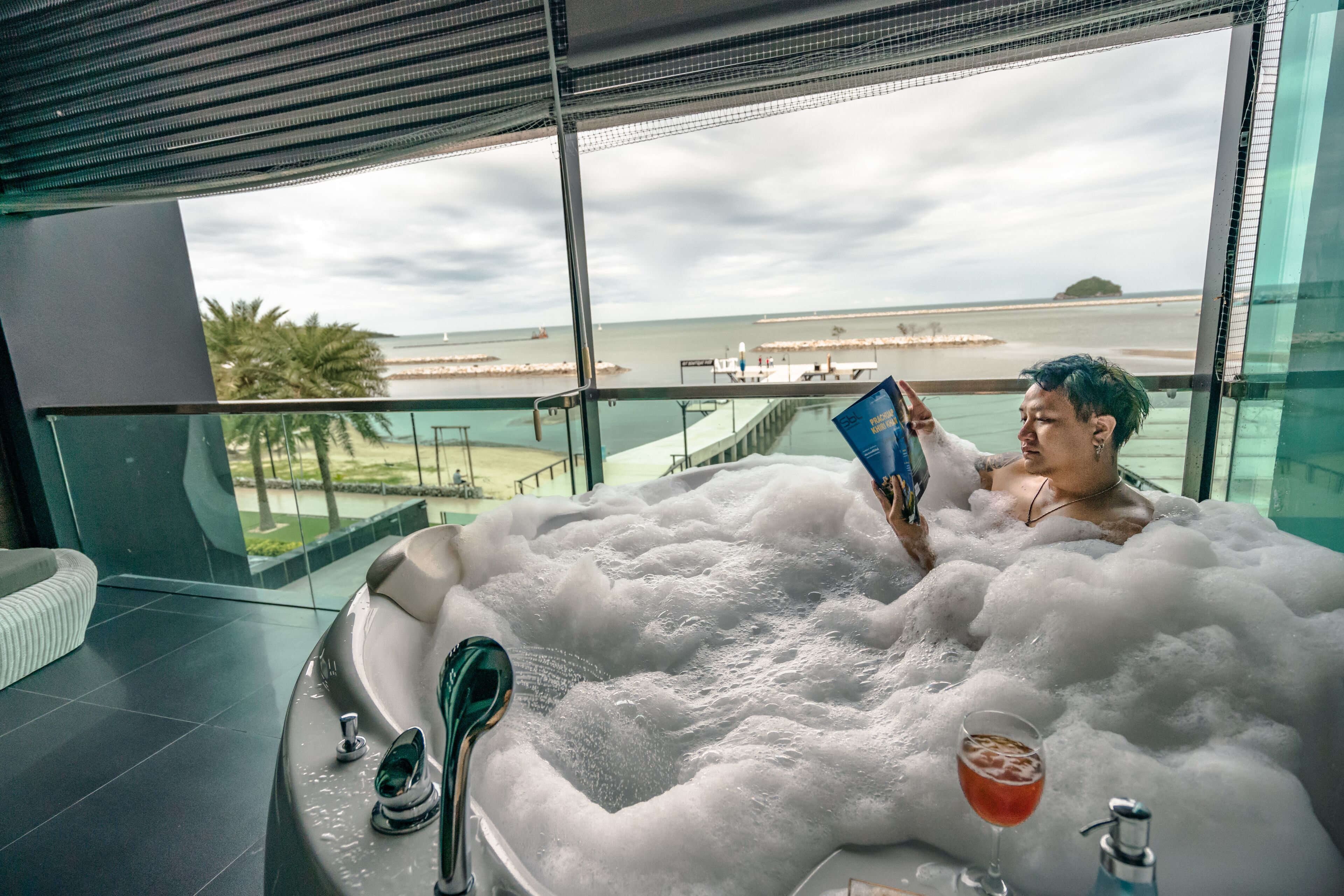 A person relaxing in a large, bubble-filled bathtub with a drink on the side, overlooking a beach and palm trees.
