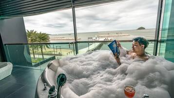 A person relaxing in a large, bubble-filled bathtub with a drink on the side, overlooking a beach and palm trees.
