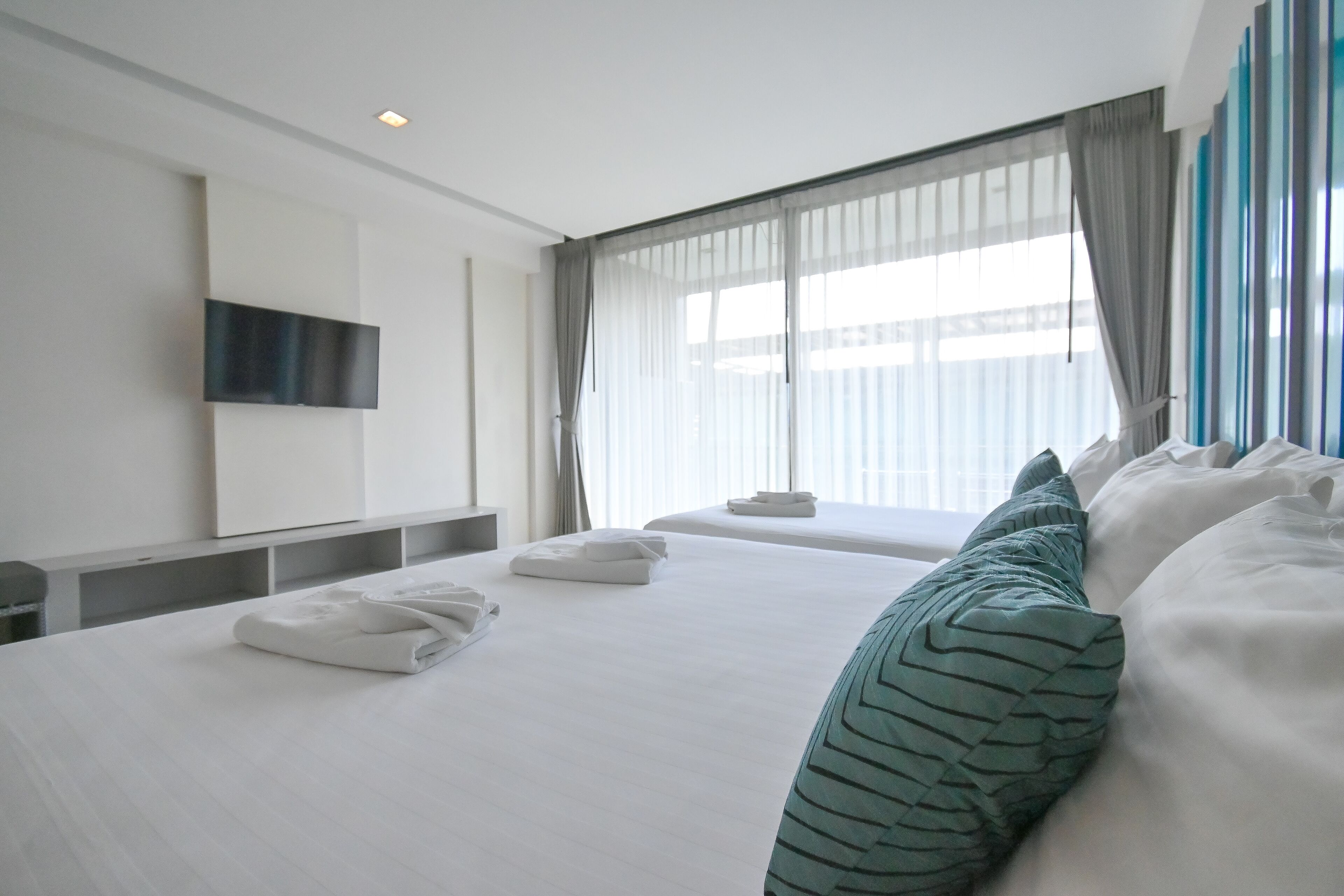 A modern hotel room with a large bed, a flat-screen TV mounted on the wall, and a large window with sheer curtains.
