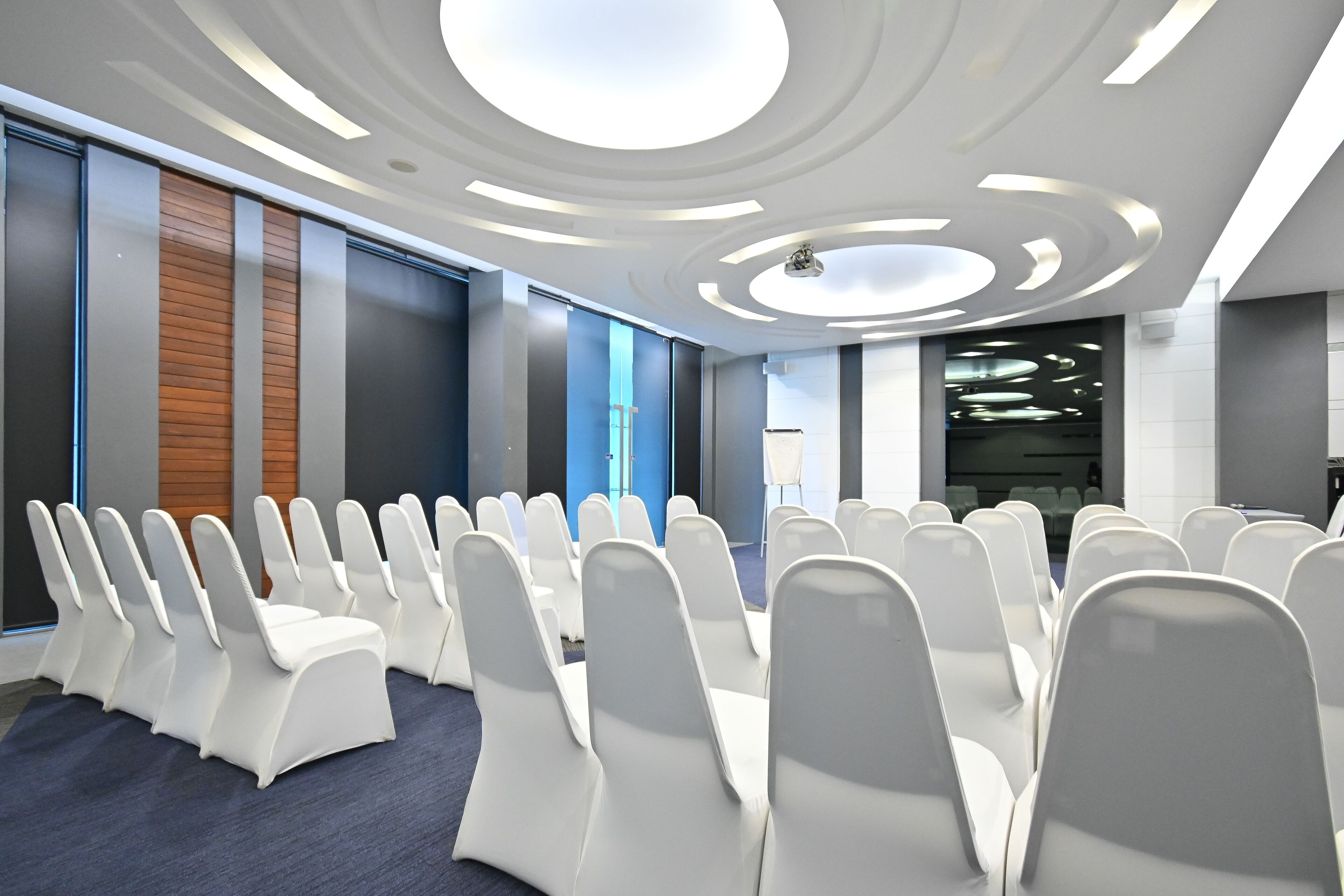 Un salón de conferencias con filas de sillas blancas, un diseño de techo moderno y un gran ventanal con vistas a la ciudad.