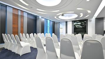Un salón de conferencias con filas de sillas blancas, un diseño de techo moderno y un gran ventanal con vistas a la ciudad.