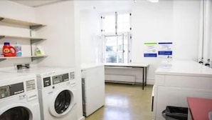A laundry room with multiple washing machines, shelves with cleaning supplies, and a window providing natural light.