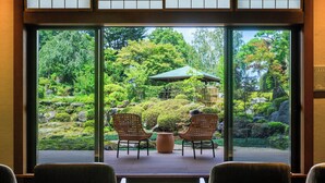 A room with a large glass window offering a view of a garden with rocks, bushes, and a gazebo. There are two wicker chairs and a small table in the foreground. - Fujisan Onsen Hotel Kaneyamaen (Fujiyoshida)