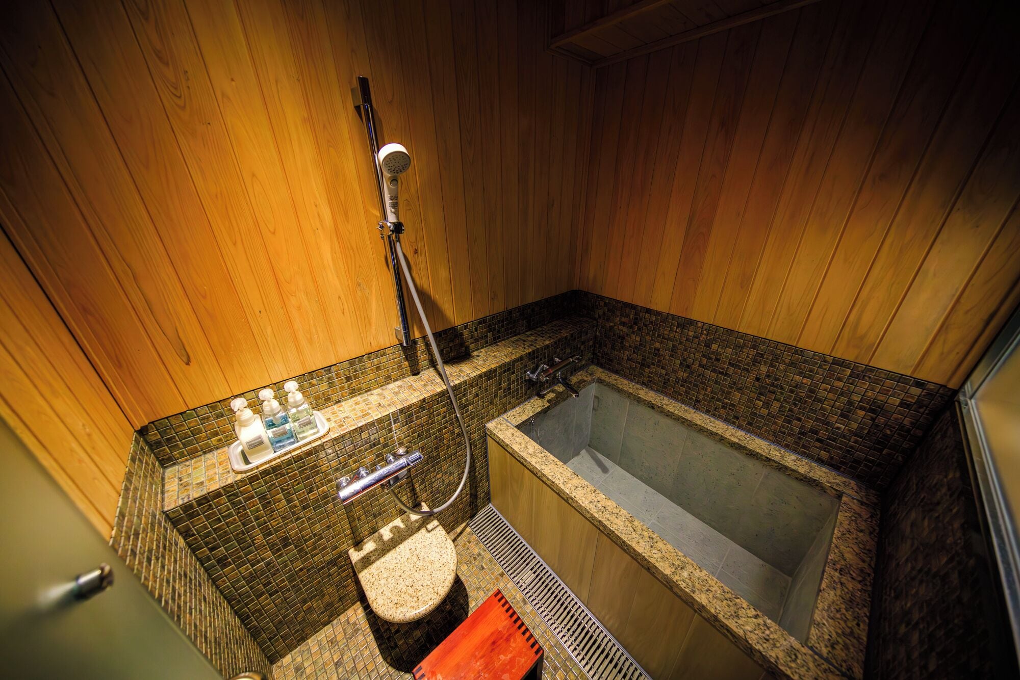 A small, wooden-finished bathroom with a single bathtub, a showerhead, and a tiled sink area.