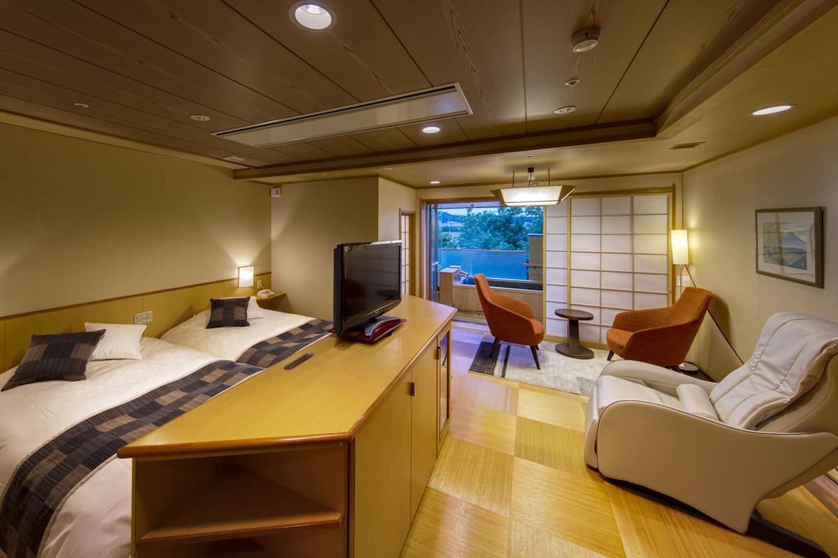 Ein Hotelzimmer mit Bett, Fernseher, Schreibtisch, zwei Stühlen und Blick ins Freie.