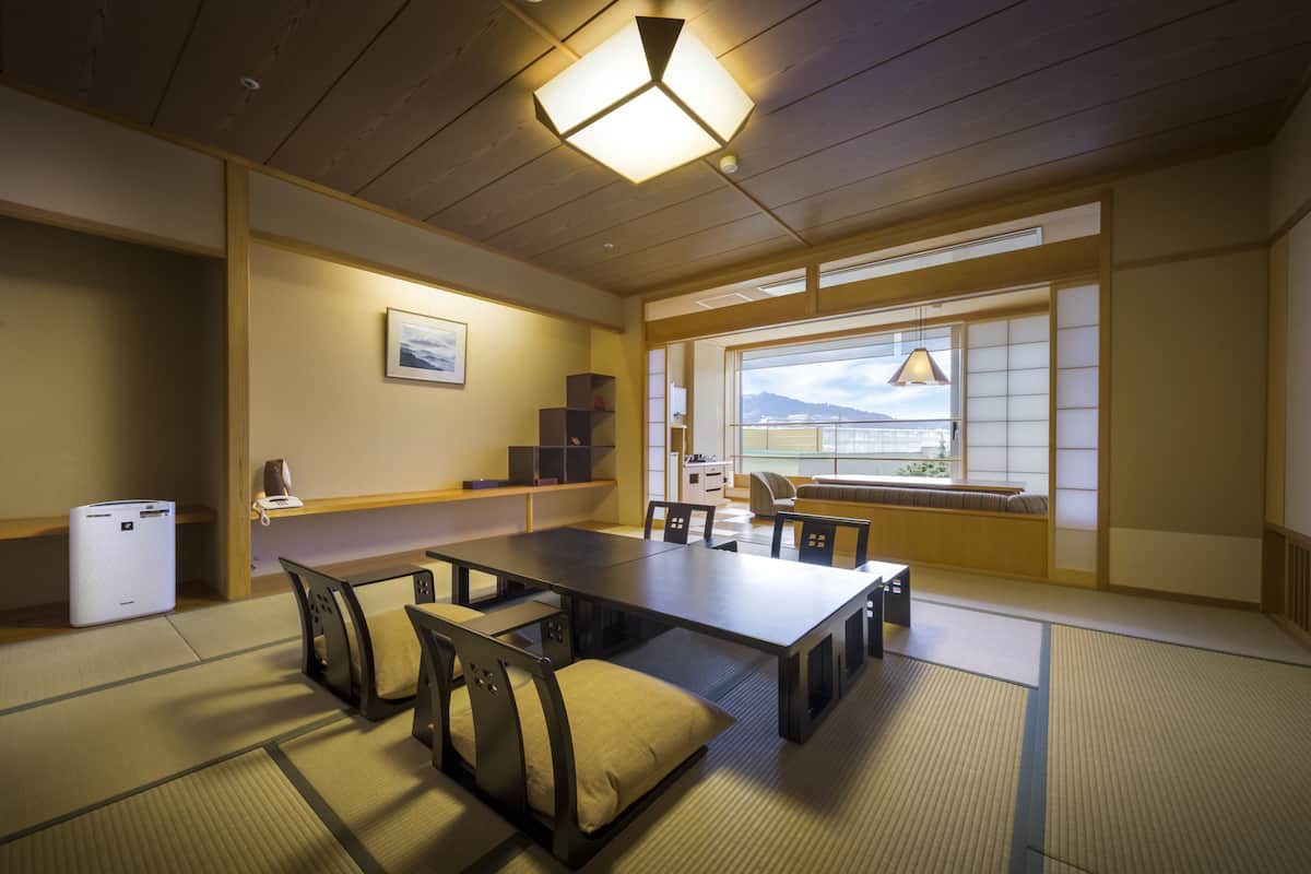 Ein traditioneller japanischer Raum mit Tatami-Boden, einem niedrigen Tisch mit Kissen, einem Schreibtisch und einem großen Fenster mit Bergblick.
