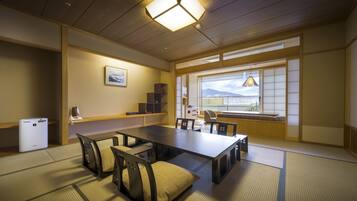 Una habitación japonesa tradicional con suelo de tatami, una mesa baja con cojines, un escritorio y un gran ventanal con vistas a las montañas.