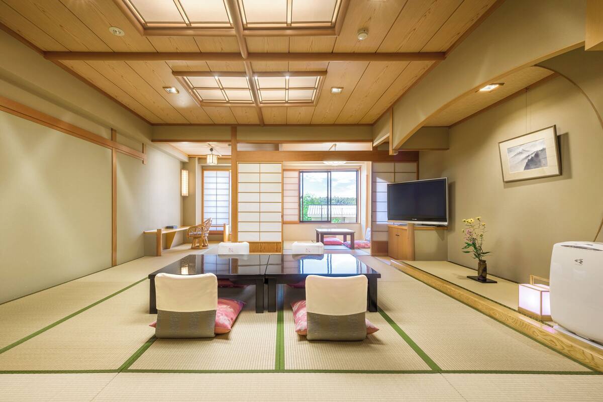 Ein traditionell gestalteter japanischer Raum mit Tatami-Böden, einem niedrigen Tisch, St ühlen, einem Fernseher und einem großen Fenster.