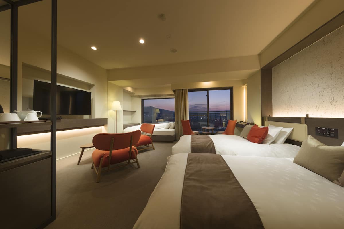 Ein Hotelzimmer mit zwei Betten, einem Schreibtisch, einem Stuhl und einem großen Fenster mit Blick auf die Stadt.