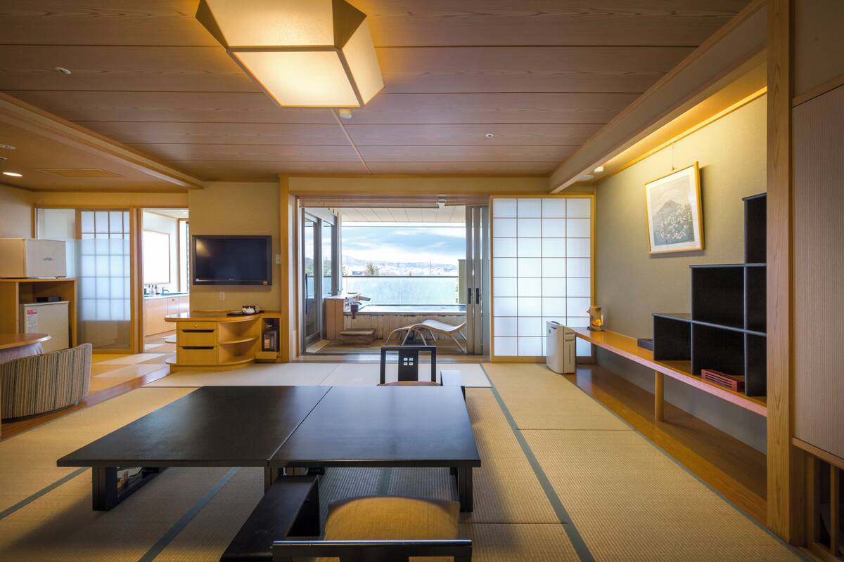 Ein traditioneller japanischer Raum mit Tatami-Bodenbelag, einem Tisch aus Tatami-Matten, einem niedrigen Holzbüro, einem Fernseher und Blick auf die Stadt durch große Fenster.