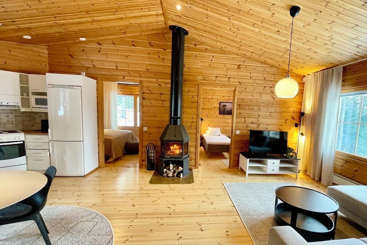 Interior acogedor de una cabaña con un estufa de leña en el centro, un comedor, una sala de estar con sofá y televisor, y una cocina con electrodomésticos modernos.